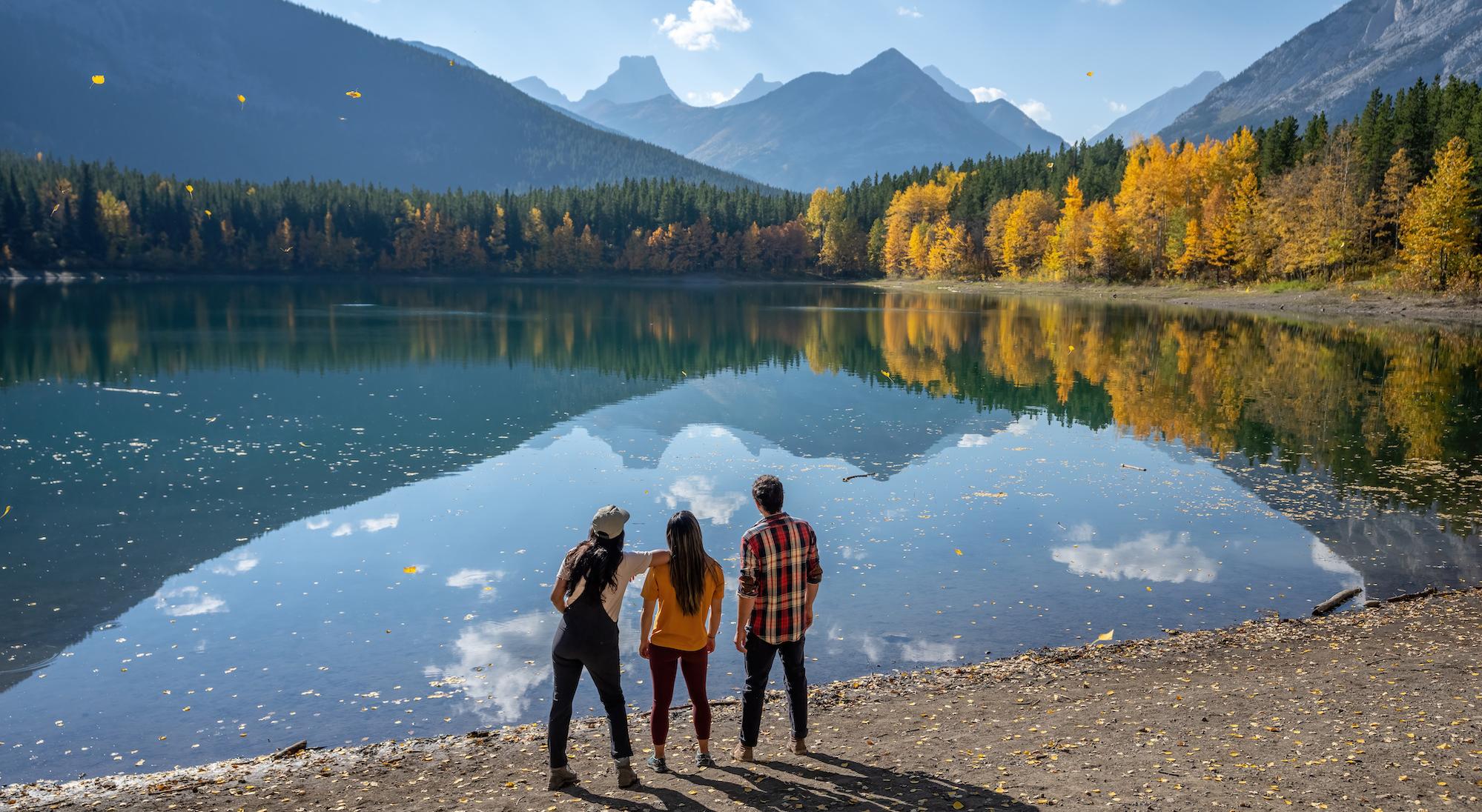 Three people standing on the edge of a lake surrounded by trees in fall colours and Rocky Mountains in the background.