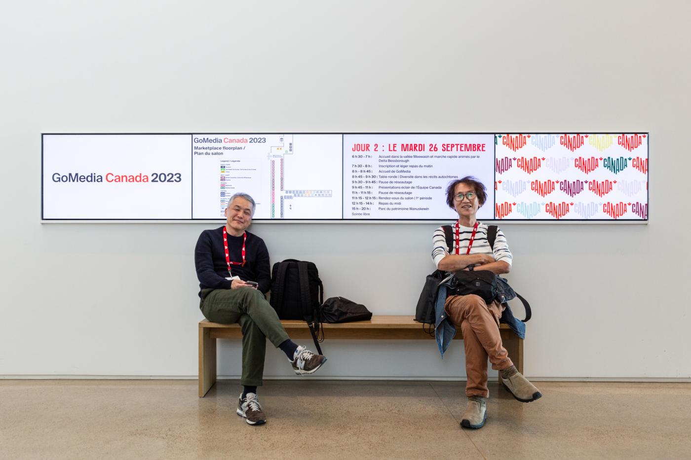 Two people sit on a bench in front of 4 side-by-side TV monitors that have branded event information on them.