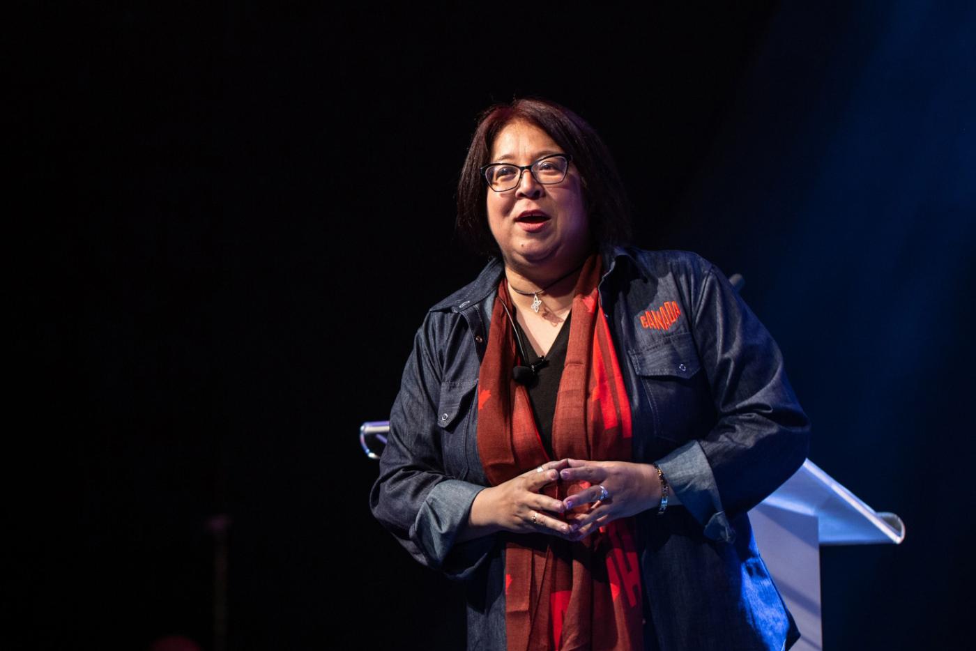 A woman is speaking at an event on stage and wearing a collared denim shirt with the Canada logo embroidered above the left breast pocket, and a red duo-toned Canada branded scarf.