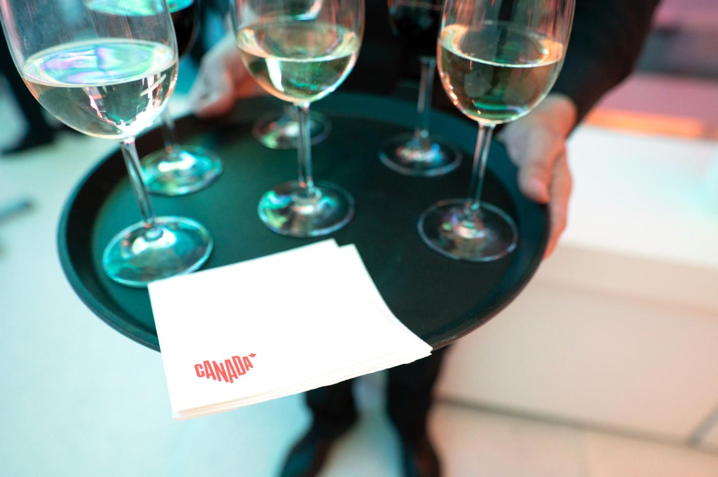 A server holds a drink tray with wine glasses and a square white napkin in the foreground which has a small red Canada logo printed in the corner of it.