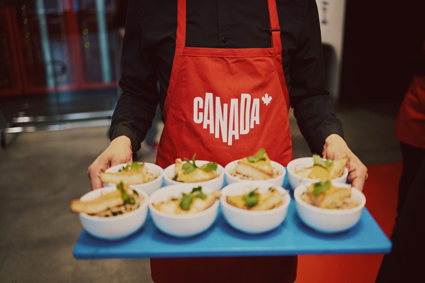 A server holds a tray of appetizers. The server is wearing a solid red apron with a white Canada logo printed in the centre