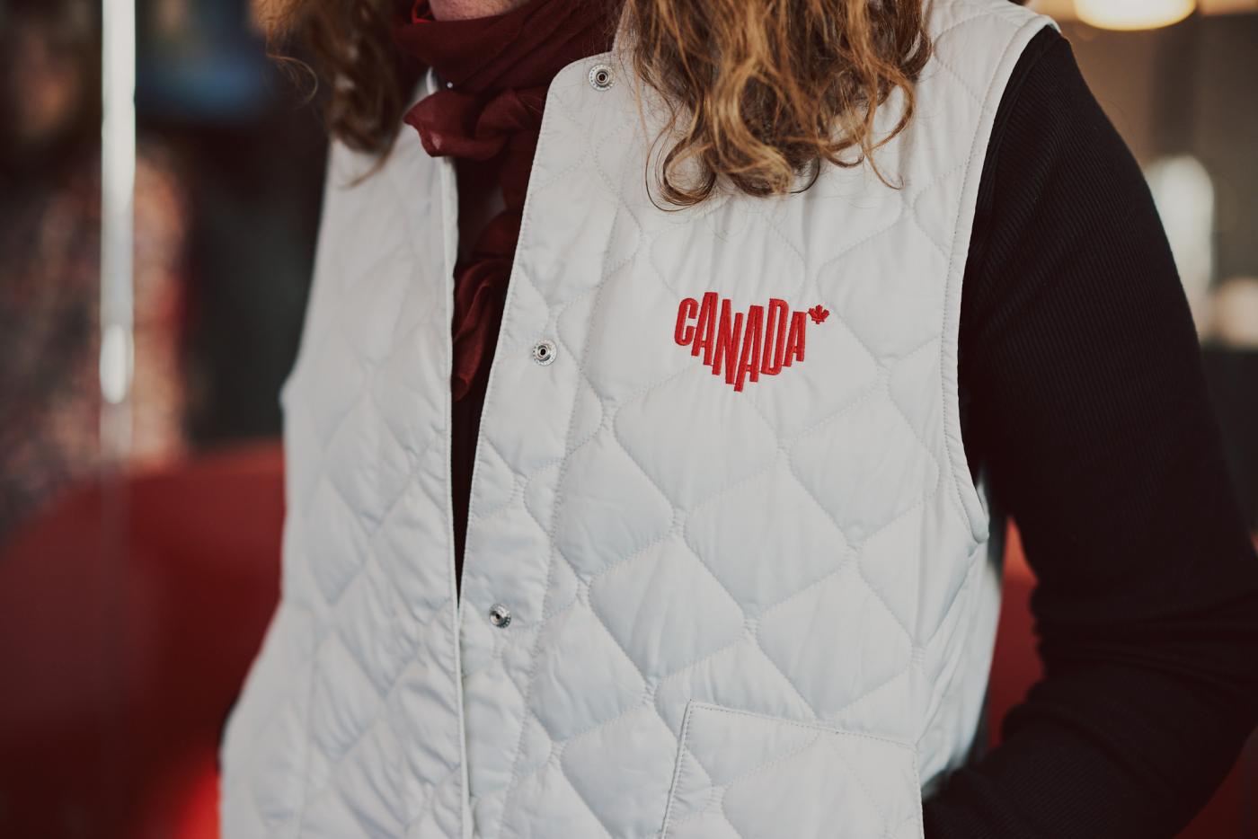 A woman stands in a white quilted vest with a red Canada logo embroidered on the front left chest area.