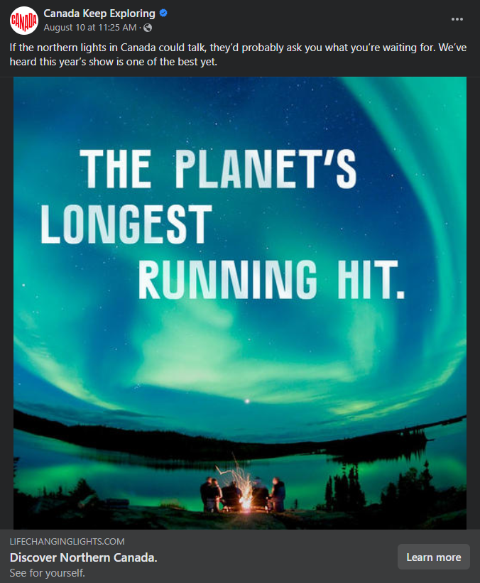 A screenshot of a social ad for Canada Keep Exploring of an image of people standing around a fire under a sky full of northern lights, with the words "THE PLANET'S LONGEST RUNNING HIT." in the sky. Caption reads: "If the northern lights in Canada could talk, they'd probably ask you what you're waiting for. We've heard this year's show is one of the best yet."