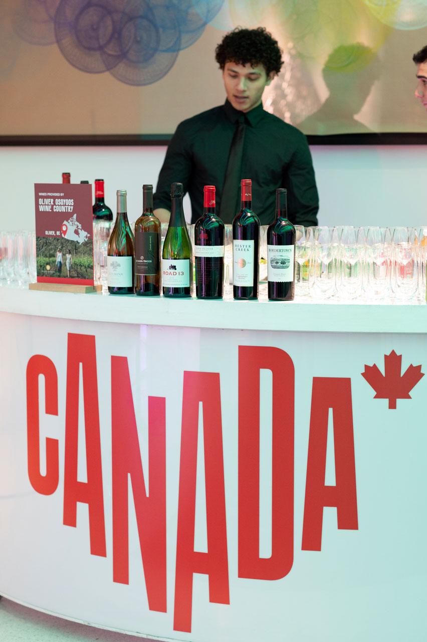 A bartender stands behind a curved white bar with a bold red Canada logo, displaying an assortment of Canadian wine bottles