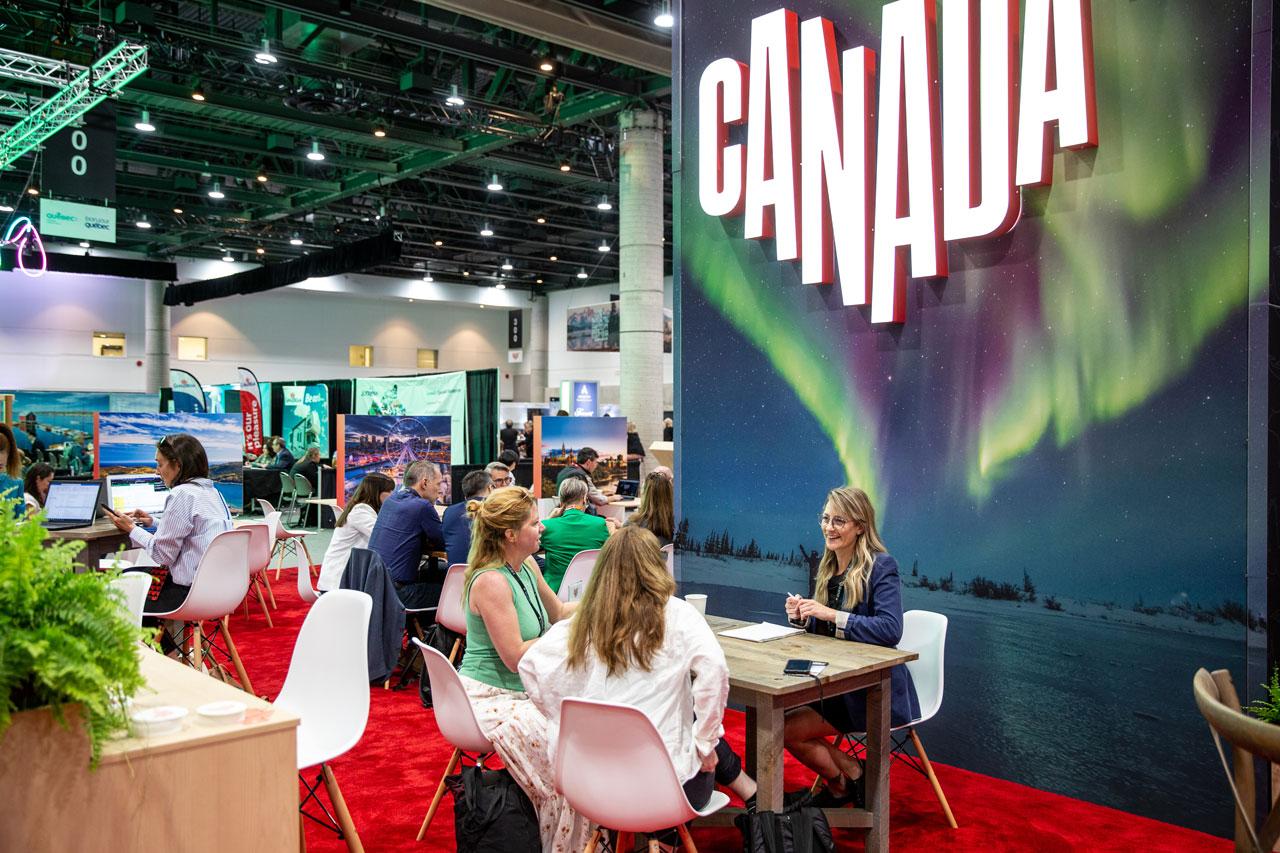 Groups of people sit at tables with a large Canada logo and aurora graphic behind them