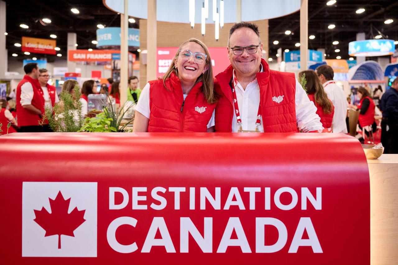 Two people standing behind a Destination Canada sign, wearing red Canada branded vests